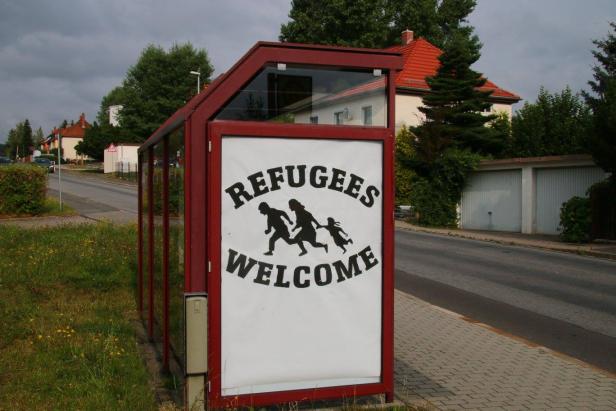 Ein rotes Bushaltestellenschild zeigt die Aufschrift „Refugees Welcome“ mit einer Familie im Laufschritt.