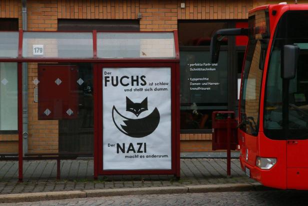 Ein roter Bus steht neben einer Bushaltestelle mit einem satirischen Plakat über Füchse und Nazis.