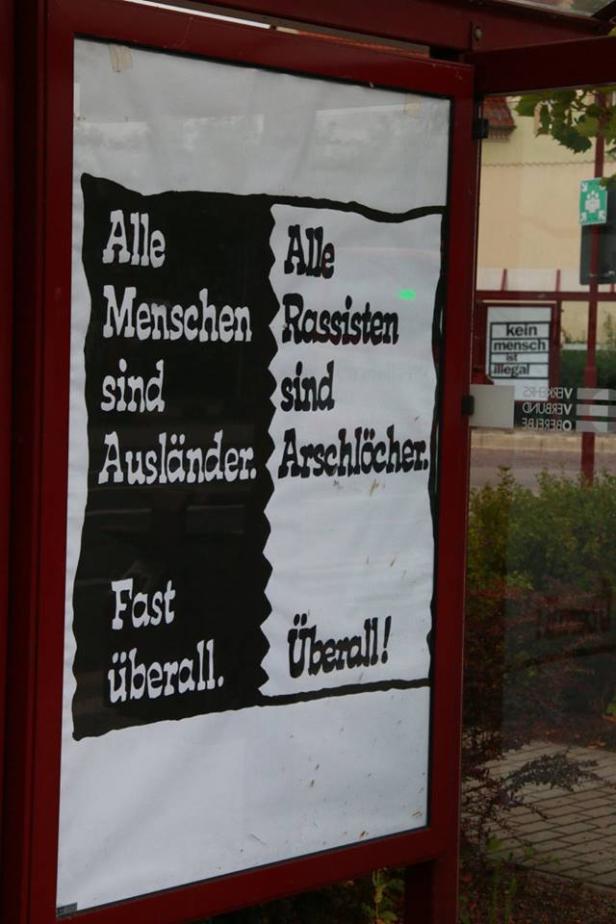 Ein Plakat in einer Bushaltestelle mit der Aufschrift „Alle Menschen sind Ausländer. Fast überall. Alle Rassisten sind Arschlöcher. Überall!“.