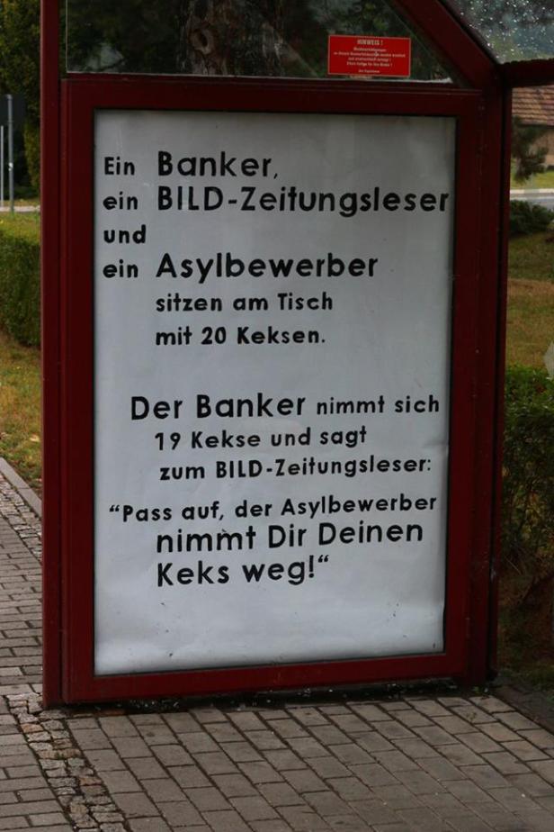 Ein Schild zeigt einen Witz über einen Banker, einen BILD-Zeitungsleser und einen Asylbewerber, die an einem Tisch mit Keksen sitzen.
