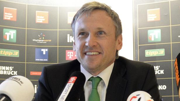 Ein Mann mit Anzug und Krawatte steht vor einer Wand mit Sponsorenlogos.