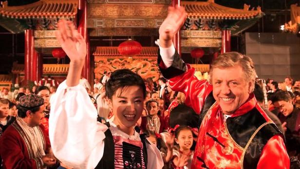 Eine Frau und ein Mann in traditioneller chinesischer Kleidung winken vor einem chinesischen Torbogen.