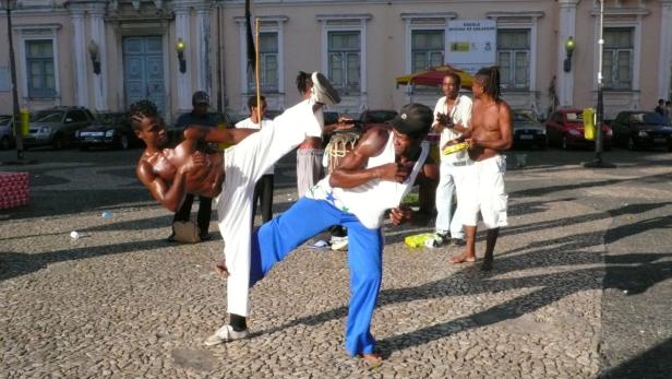 Zwei Männer führen auf einem Platz Capoeira vor, begleitet von Musikern.