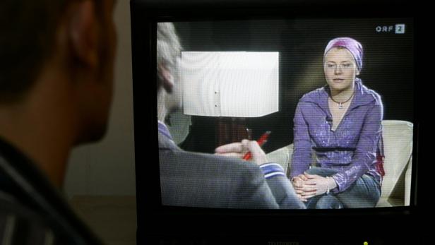 Ein Mann sieht fern; im Fernsehen ist eine Frau zu sehen, die interviewt wird.
