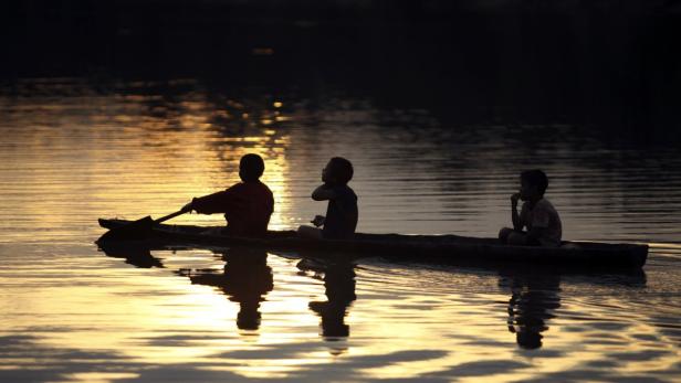 Drei Kinder fahren in einem kleinen Boot auf einem ruhigen Gewässer in der Dämmerung.