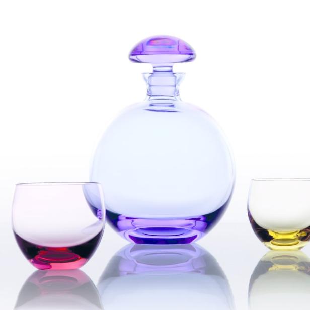 Ein lilafarbener Dekanter aus Glas steht neben zwei farbigen Gläsern.