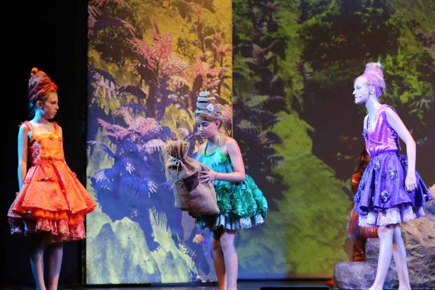 Drei junge Schauspielerinnen in farbenfrohen Kostümen stehen auf einer Bühne vor einer Waldkulisse.