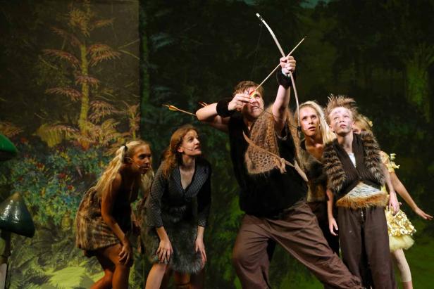 Eine Gruppe von Schauspielern auf einer Bühne mit Waldkulisse, ein Mann zielt mit Pfeil und Bogen.