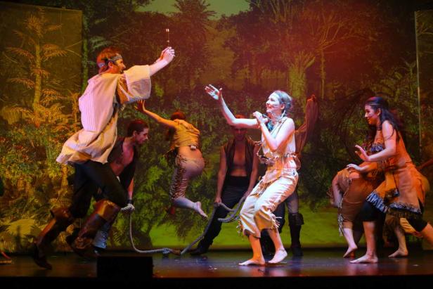 Eine Theaterszene mit mehreren Darstellern in Kostümen vor einem Waldhintergrund.