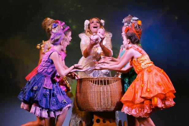 Eine Frau hält eine Puppe über einen Korb, umgeben von Mädchen in bunten Kleidern auf einer Bühne.
