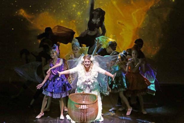 Eine Frau in einem weißen Kleid steht mit ausgebreiteten Armen auf einer Bühne, umgeben von Tänzern.