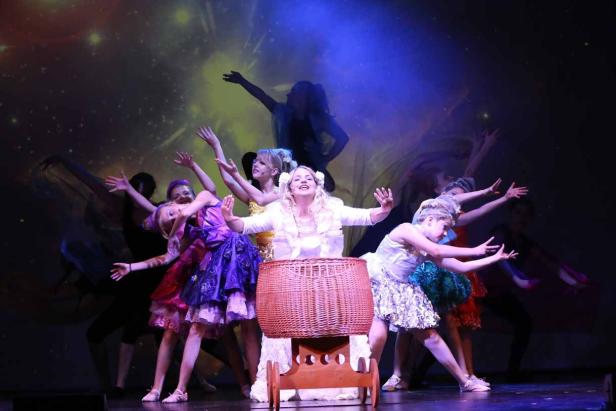 Eine Frau in Weiß steht vor einem Korbwagen, umgeben von Tänzern auf einer Bühne.