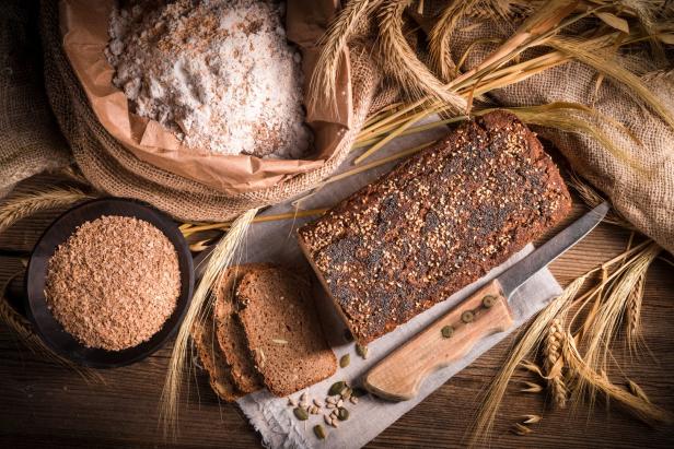Ein Laib Brot mit Saaten, Mehl und Getreideähren auf einem Holztisch.