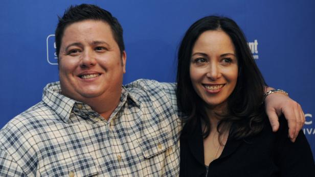 Ein Mann im karierten Hemd und eine Frau lächeln vor einem blauen Hintergrund.