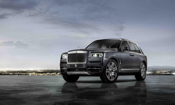 Ein grauer Rolls-Royce Cullinan vor einer Stadtlandschaft im Abendlicht.