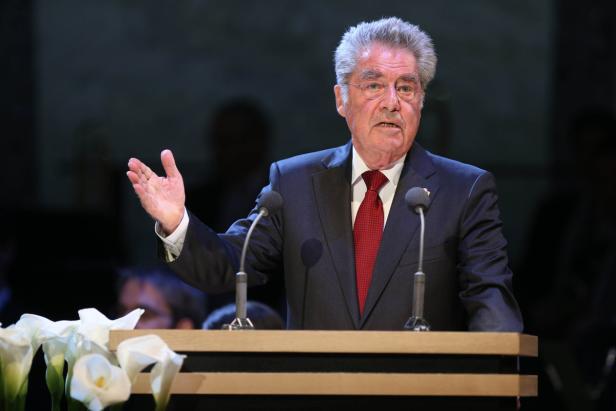 Ein älterer Mann mit Anzug und roter Krawatte gestikuliert an einem Rednerpult.