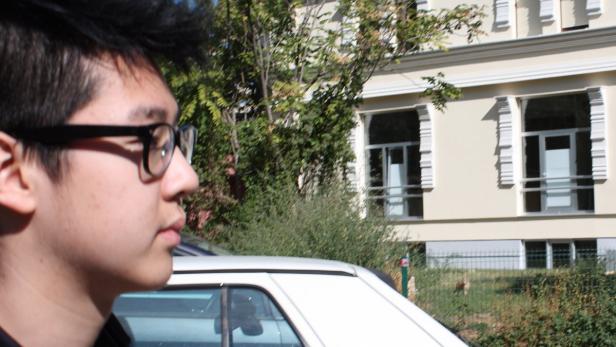 Ein junger Mann mit Brille steht vor einem Gebäude und einem weißen Auto.