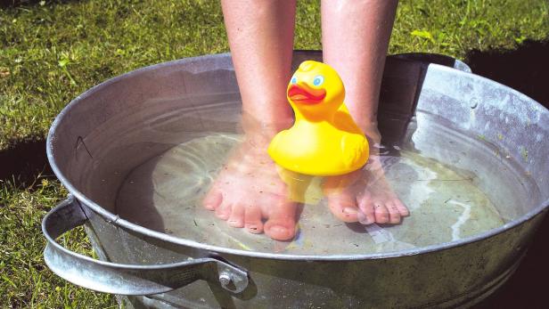 Füße stehen mit einem gelben Gummiente in einer Zinkwanne mit Wasser.