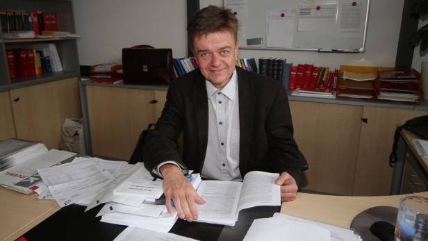 Ein Mann sitzt an einem Schreibtisch voller Papiere und liest in einem Buch.