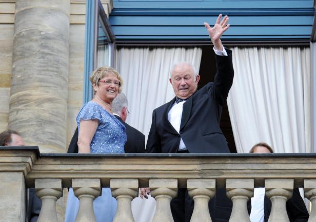 Ein Mann im Smoking winkt von einem Balkon, neben ihm eine Frau in einem blauen Kleid.