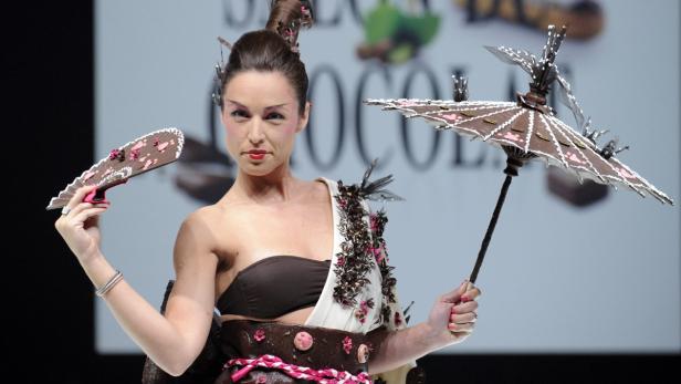 Eine Frau in einem Schokoladenkleid mit Fächer und Schirm auf einer Modenschau.