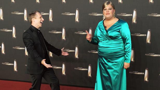 Ein Mann im Anzug und eine Frau im türkisfarbenen Kleid posieren vor einer Wand mit der Aufschrift „Deutscher Fernsehpreis“.