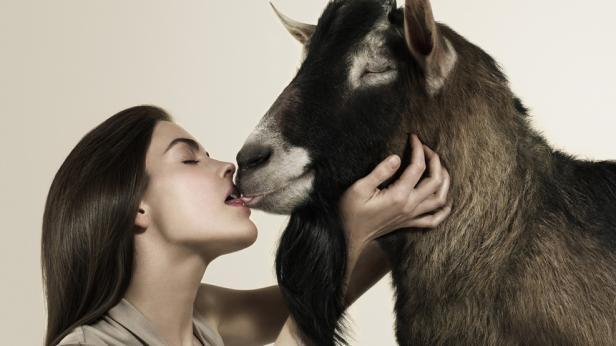Eine Frau leckt das Maul einer braunen Ziege.