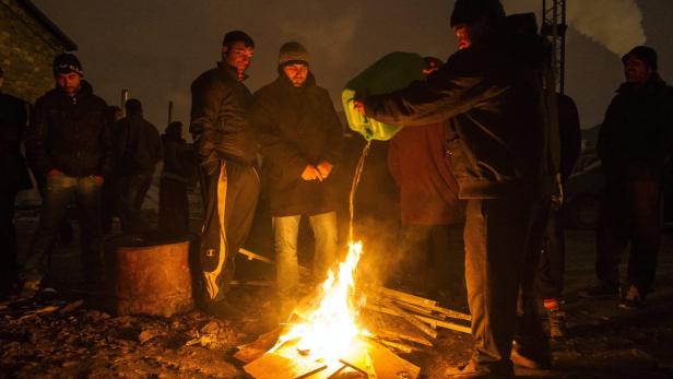 Eine Gruppe von Männern wärmt sich an einem Lagerfeuer, das mit einer Flüssigkeit aus einem Kanister entzündet wird.