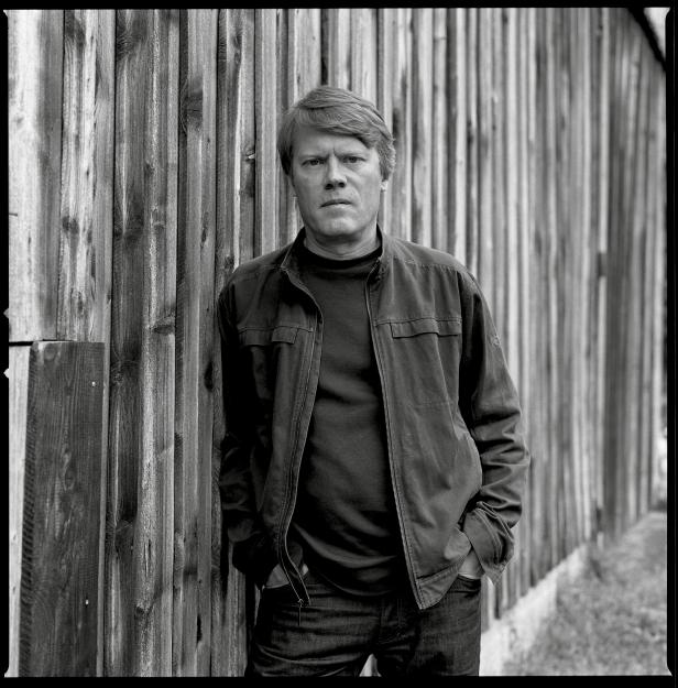 Ein Mann in Jacke und Jeans steht vor einer Holzwand.