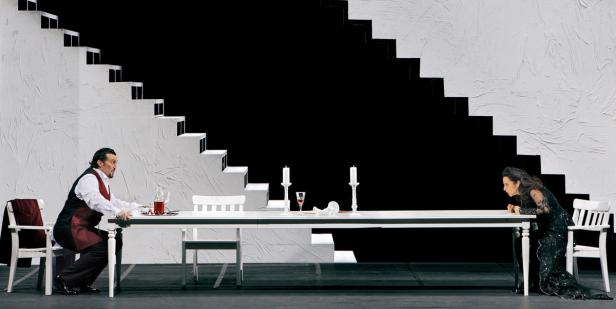 Eine Szene aus einer Opernaufführung mit zwei Sängern an einem langen Tisch vor einer Treppe.