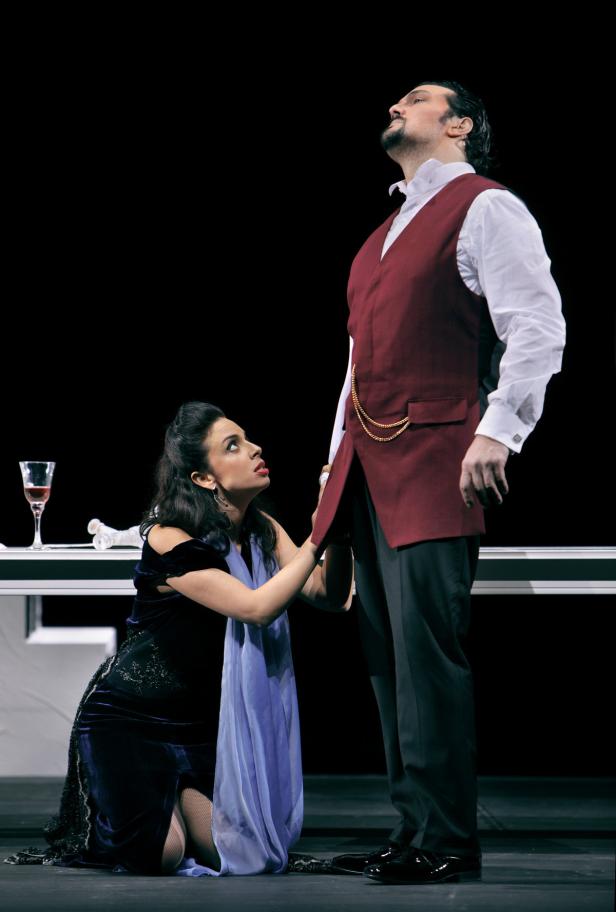 Eine Frau kniet vor einem Mann in einem roten Samtweste auf einer Bühne.