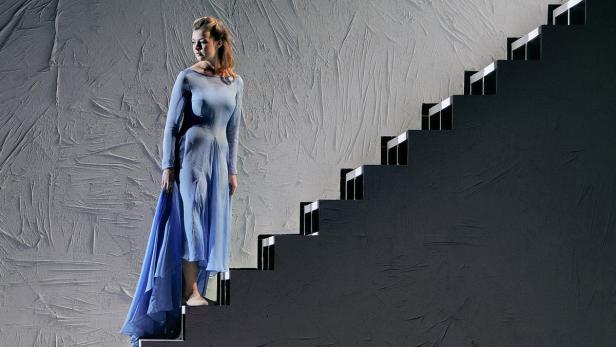 Eine Frau in einem hellblauen Kleid steht auf einer Treppe vor einer strukturierten Wand.