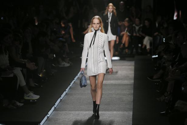 Eine blonde Frau läuft in einem weißen Kleid und schwarzen Stiefeln über einen Laufsteg.