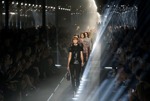 Eine Modenschau mit Models, die den Laufsteg entlanggehen, während Zuschauer applaudieren.