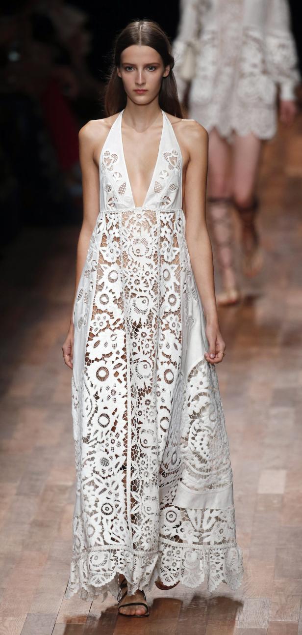 Ein Model präsentiert ein langes, weißes Spitzenkleid auf dem Laufsteg.