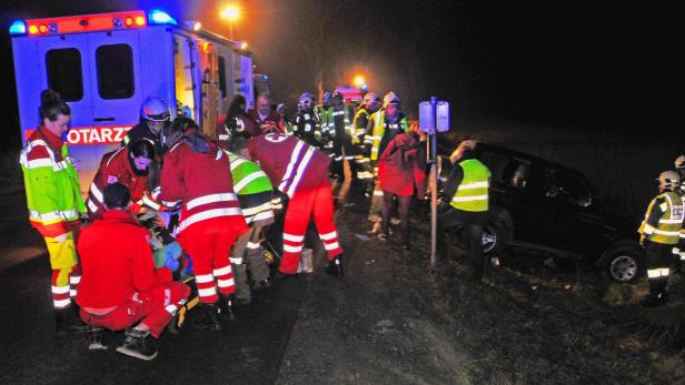 Rettungskräfte versorgen eine verletzte Person neben einem Rettungswagen und einem verunglückten Auto.