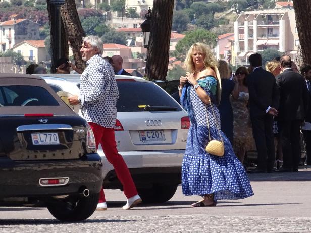 Eine Frau in einem blauen Kleid und ein Mann in roter Hose gehen an Autos vorbei.