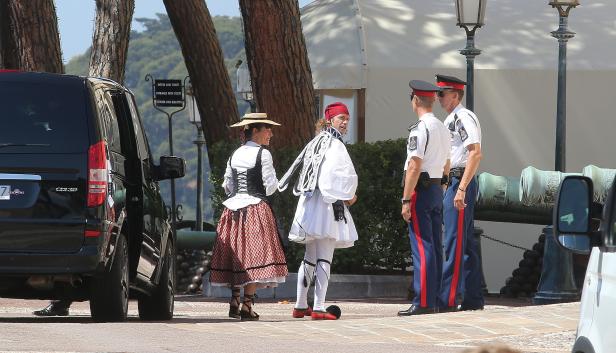 Eine Frau in Tracht und ein Mann in traditioneller griechischer Kleidung stehen neben zwei Wachen.