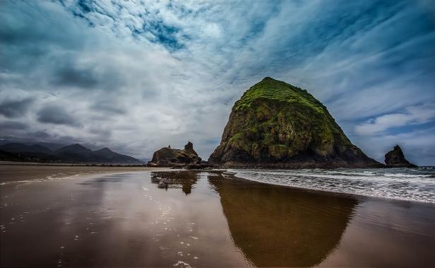 Ein großer, grüner Felsen liegt an einem Strand unter einem bewölkten Himmel.