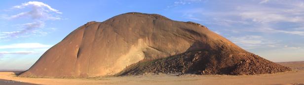 Ein großer, glatter Felsen erhebt sich in einer Wüstenlandschaft unter einem blauen Himmel.