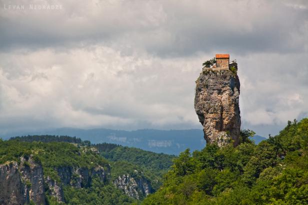 Ein kleines Haus mit rotem Dach steht auf einem hohen Felsen inmitten grüner Hügel.