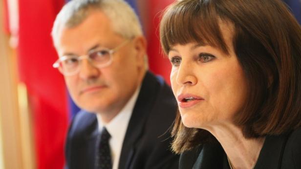 Eine Frau spricht, während ein Mann mit Brille im Hintergrund sitzt.