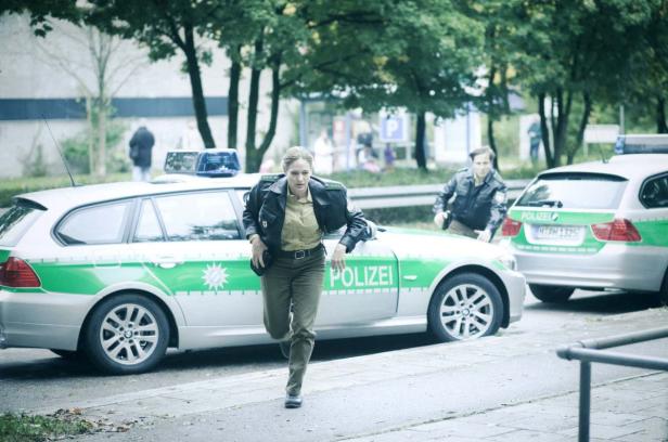 Eine Polizistin rennt vor einem Streifenwagen, während ein Polizist folgt.