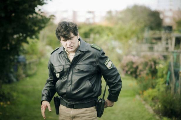 Ein Polizist in Lederjacke mit blutiger Hand geht durch einen Garten.