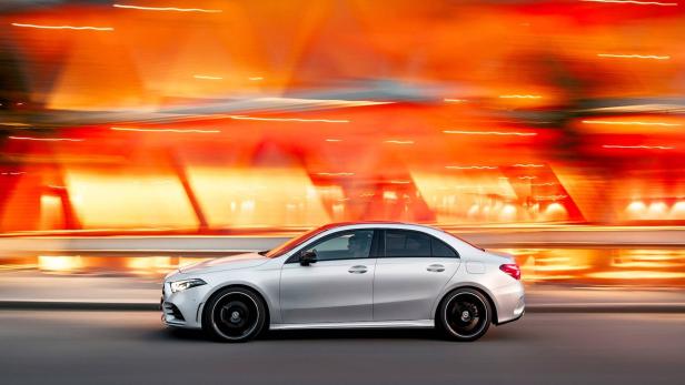 Eine silberne Mercedes-Benz A-Klasse Limousine fährt auf einer Straße mit verwischtem, orangefarbenem Hintergrund.