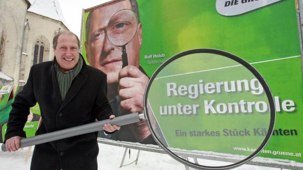 Rolf Holub, Spitzenkandidat der Grünen Kärnten, posiert vor einem Wahlplakat mit dem Slogan „Regierung unter Kontrolle“.