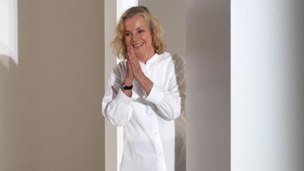 Eine lächelnde Frau mit blonden Haaren und weißem Hemd steht vor einer Wand.