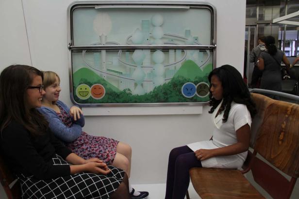 Drei Frauen sitzen auf einer Bank vor einer Grafik mit Smileys und einer stilisierten Stadtlandschaft.