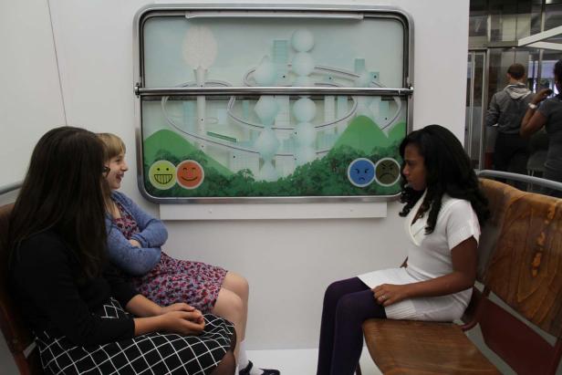 Drei junge Frauen sitzen in einem Raum mit einer Grafik, die Emotionen darstellt.