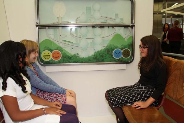 Drei Mädchen sitzen vor einer Grafik mit stilisierten Gesichtern und einer Landschaft.